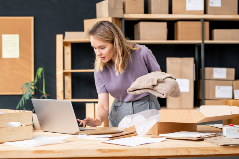 Pretty young female manager of online shop with folded sweater using laptop